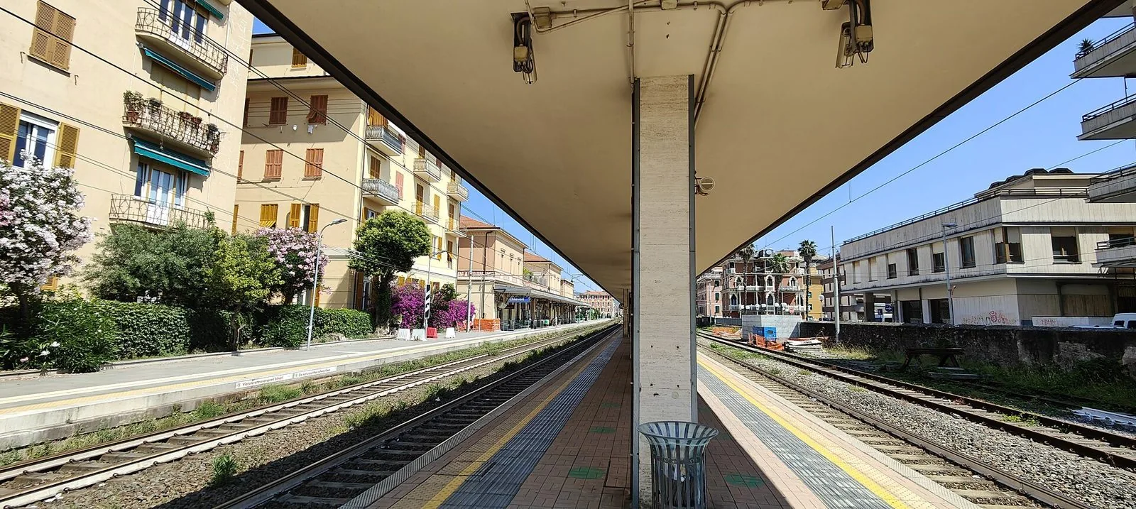 Binari e pensilina della stazione ferroviaria di Chiavari tra palazzi color pastello e oleandri in fiore